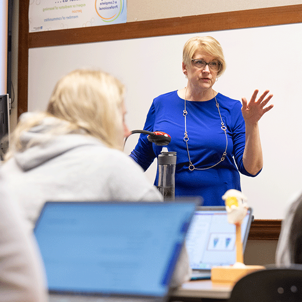 A white female professor stands at the front of a classroom. She has short blond hair, and she wears glasses, a long-sleeved blue dress, and a long gold necklace. A white female student with blond hair, wearing a white sweatshirt, sits in front of the professor. Laptops are on the desks. A whiteboard is in the background.