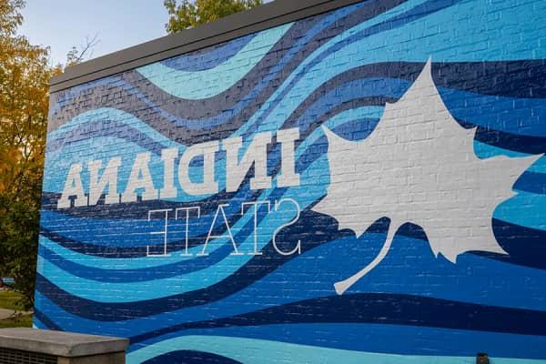 Large mural with a white Sycamore leaf and “Indiana State” written in white text with various shades of wavy blue lines as the background on the left side of the image while on the right is a black lamp post with the light on with trees with yellow fall leaves behind. 