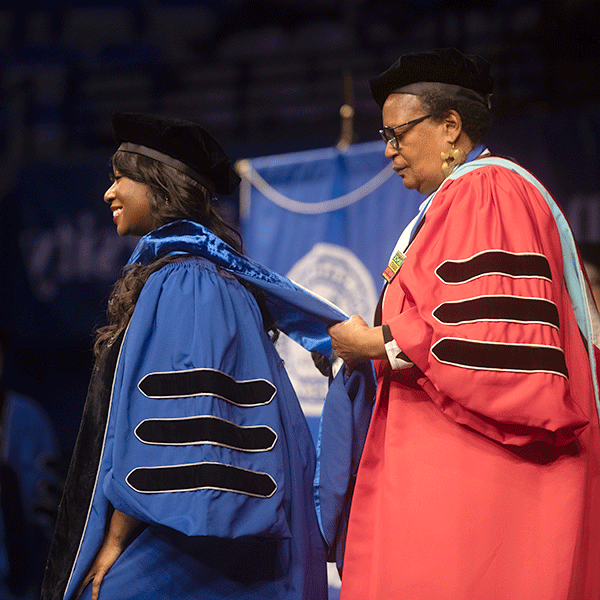 A Black female professor wearing a red commencement robe, places doctoral hood over the head of a Black female student during a commencement ceremony for Graduate Students.